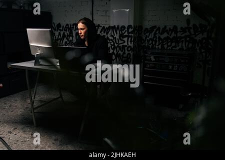 Un homme hacker concentré dans un sweat-shirt noir utilisant un ordinateur portable et un ordinateur de bureau pour pirater le système réseau de la pièce sombre avec mur de briques. Banque D'Images