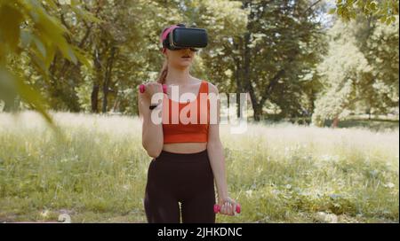 Une fille sportive dans un casque VR fait des exercices de fitness avec des haltères à l'extérieur dans un parc ensoleillé. Entraînement en ligne. Jeune femme en sportswear regardant la réalité virtuelle 3D vidéo sport bodybuilder jeu Banque D'Images