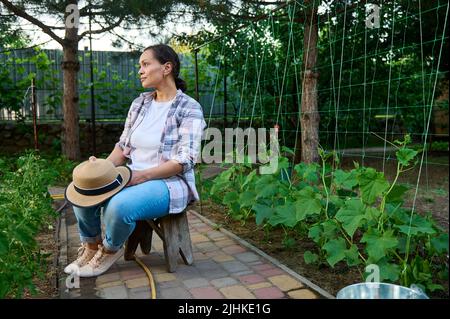 Le jardinier féminin, repose après le travail à la ferme, s'assoit sur des tabouret entre des rangées de semis de tomates et de concombres. Banque D'Images