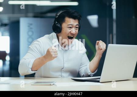 Un homme d'affaires asiatique heureux regardant un match sportif en ligne, un homme au bureau regardant l'écran d'un ordinateur portable applaudisse Banque D'Images