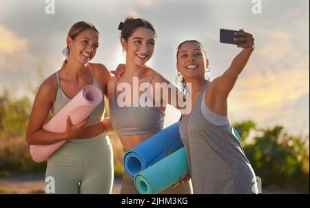 Belles femmes de yoga utilisant un téléphone portable pour prendre des selfies tout en tenant des tapis de yoga dans la pratique en plein air dans la nature éloignée. Groupe diversifié de jeunes souriants Banque D'Images