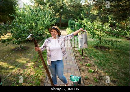 Joyeuse femme jardinier, fermier, horticulteur aime le jardinage au début du printemps, se tient dans une ferme écologique avec une houe, ripper et arrosoir dedans Banque D'Images