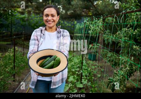 Belle femme horticultrice afro-américaine, sourit un sourire enragé, regardant l'appareil photo, tenant un chapeau de paille rempli de mûres fraîchement cueillies prêt-à-manger Banque D'Images