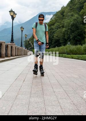 jeune homme en vêtements bleus en équipement de protection, sur des patins à roulettes le long d'un remblai sur fond de forêt et de montagnes en été Banque D'Images