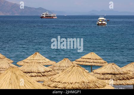 Plage ensoleillée avec parasols en osier, station d'été. Vue sur la mer Méditerranée, les bateaux à moteur et les montagnes dans la brume Banque D'Images