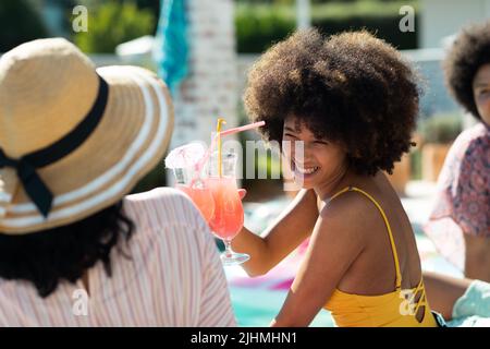 Jeune femme biraciale souriante avec un cocktail de toasting de cheveux afro avec un ami tout en appréciant la fête de la piscine Banque D'Images