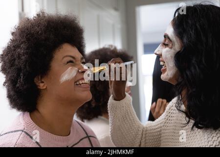 Bonne jeune femme biracial avec de la crème de beauté sur le visage appliquant le paquet de visage à l'amie femelle à la maison Banque D'Images