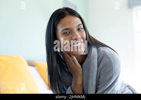 Portrait d'une jeune femme biraciale heureuse avec cheveux longs droits portant un peignoir contre le mur à la maison Banque D'Images