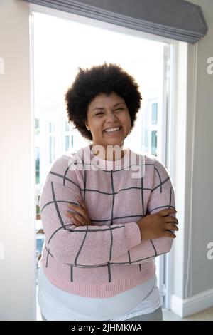 Portrait de jeune femme biracial souriante avec cheveux afro et bras croisés debout contre la fenêtre Banque D'Images