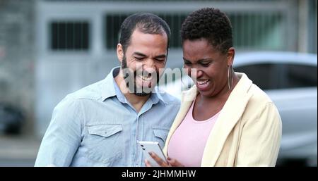 Couple interracial riant dans la rue tenant le téléphone cellulaire. Relation mixte et franche avec un smartphone. La vraie vie rire et sourire Banque D'Images