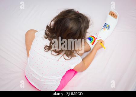 Une petite fille avec une jambe cassée à la maison sur le lit dessine avec des stylos feutre sur une fonte Banque D'Images