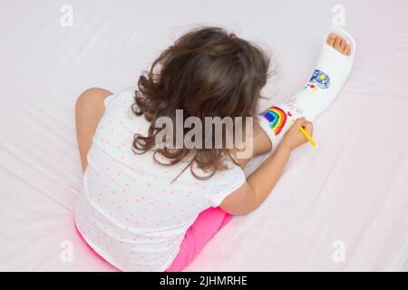 Une petite fille méconnaissable avec une jambe cassée à la maison sur le lit dessine avec des stylos feutre sur une fonte orthopédique. Comment s'amuser avec une jambe cassée Banque D'Images