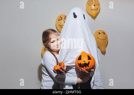 Joyeux Halloween. Une fille embrasse le fantôme et donne de la citrouille orange. Enfant en costume blanc. Boules jaunes peintes. Différentes émotions de joie, de colère, de rire. Design festif, concept de fête. Mise au point sélective douce Banque D'Images