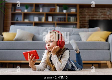 Surprise petite fille portant des écouteurs rouges couchés sur la moquette à la maison. Jolie fille tenant un smartphone, regardant des dessins animés, des messages texte, la navigation Banque D'Images