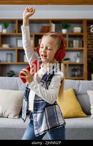 Bonne petite fille portant un casque rouge dansant et chantant à l'aide d'un smartphone comme un microphone. Enfant mignon Profitez de votre musique préférée Banque D'Images