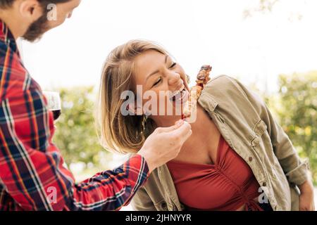 Une jeune femme curvy rit alors qu'elle mange des brochettes de viande des mains de son ami lors d'un voyage à la campagne. Les jeunes adultes se sont amusés Banque D'Images