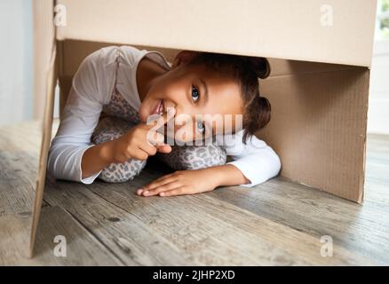 Portrait d'une petite fille hispanique mignonne jouant avec une boîte en carton dans un nouvel appartement. Jolie fille de race mixte se cachant dans une boîte et souriant dans une maison Banque D'Images