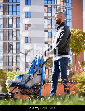 Le père passe du temps avec un nouveau-né à l'extérieur. Jeune homme barbu marchant avec bébé poussette dans la rue dans la cour moderne des immeubles résidentiels de la ville. Banque D'Images