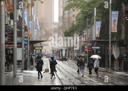 Sydney, Australie - 19 juillet 2022. Des piétons portant des parasols traversent des voies ferrées légères sur George Street dans le centre-ville de Sydney. Banque D'Images