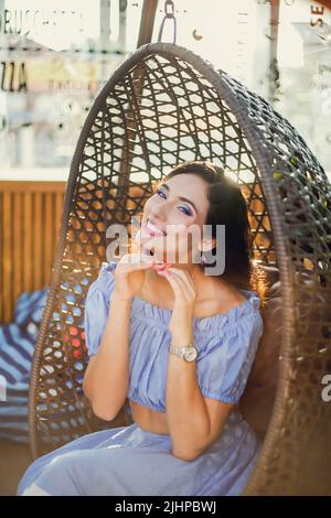 Une belle jeune femme est assise dans une chaise suspendue en osier sur la terrasse d'été d'un café de la ville, elle a soulevé ses mains au menton, regardant la caméra Banque D'Images