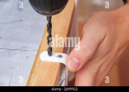 Un homme fore des trous dans un crochet en fer avec une perceuse pour accrocher un écran en bois sur une batterie. Banque D'Images