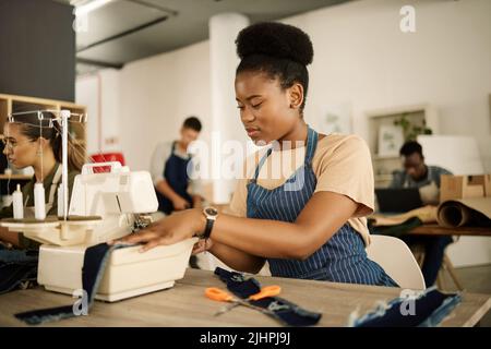 Couture d'un jeune designer sur une machine. Un Jean sur mesure afro-américain est surpiqué. Designer de mode travaillant à sa table. Couture de couturière Banque D'Images
