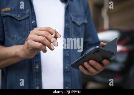Gros plan. Dépendance malsaine fumer cigaret homme de lire les médias sociaux en utilisant son smartphone portant un Jean chemise et un t-shirt blanc debout à l'extérieur sur fond urbain ville. Aucune face visible. Banque D'Images
