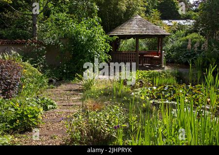 Jardin anglais avec étang ornemental avec élément de maison d'été et chemin de gravier avec des dormeuses en bois, Angleterre Banque D'Images