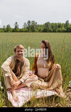 Homme souriant assis sur une couverture près de la femme enceinte dans le champ Banque D'Images