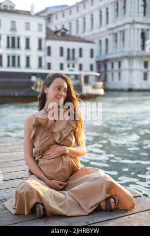 Femme enceinte souriante assise sur une jetée en bois près de la rivière à Venise Banque D'Images