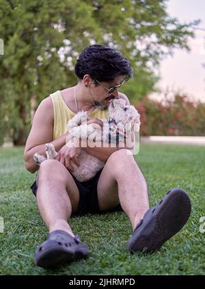 latino brun avec des lunettes de soleil assis sur la pelouse de son jardin jouant avec son animal de compagnie blanc jouet paodle un après-midi d'été. Mise au point sélective. Verti Banque D'Images