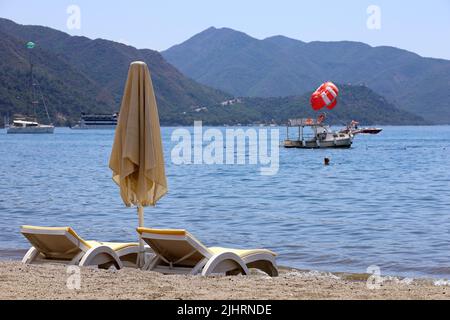 Vider les chaises longues et le parasol sur une plage. Vue sur la mer Méditerranée, bateau avec parachute ascensionnel et montagnes verdoyantes en brume Banque D'Images