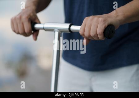 Gros plan homme mains tenant la poignée de scooter dans les rues ou se garer après le travail à l'extérieur, portant un t-shirt bleu foncé et un short clair. Aucune face visible. Mise au point sélective sur la gauche. Banque D'Images