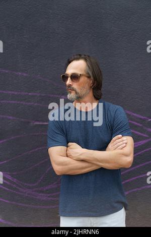 Beau homme d'âge moyen debout avec les mains pliées à côté du mur urbain foncé portant des lunettes de soleil, un t-shirt bleu foncé et un short clair. Homme d'affaires ou indépendant à l'extérieur. Banque D'Images