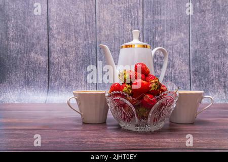 Vase en verre rempli de fraises rouges mûres sur table en bois. À proximité se trouve une théière en porcelaine et deux tasses. Banque D'Images