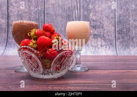 Vase en cristal rempli de fraises rouges mûres sur une table en bois. À proximité se trouvent des verres à vin remplis de lait et de cocktails au chocolat. Banque D'Images