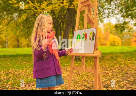 petite fille artiste avec un pinceau et des peintures dans ses mains en automne dans le parc dessine un paysage avec des feuilles sur toile Banque D'Images