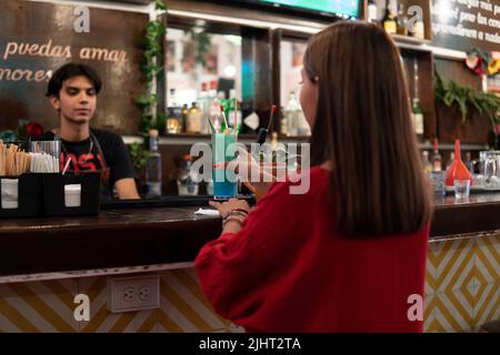 Une fille aux cheveux bruns est à un bar demandant au barman une boisson. Un barman sert une boisson bleue à une fille latine dans une robe rouge. Elle fille dans un club qui demande une boisson alcoolisée. Banque D'Images