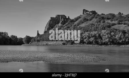 Vue en noir et blanc sur les ruines du château de Devín au-dessus du Danube en Slovaquie, avec une eau calme et un paysage historique. Banque D'Images