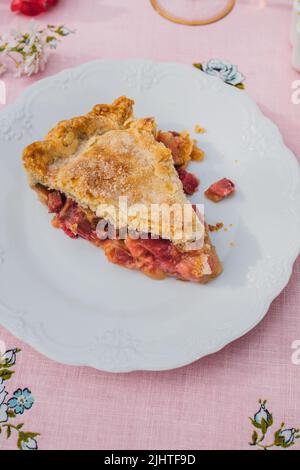 une seule tranche de tarte à la rhubarbe sur plaque blanche, nappe rose avec roses bleues Banque D'Images