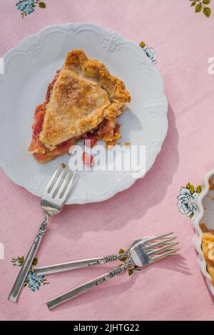 une seule tranche de tarte à la rhubarbe sur plaque blanche, nappe rose avec roses bleues Banque D'Images