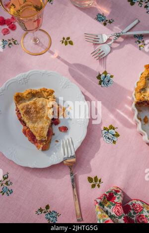 une seule tranche de tarte à la rhubarbe sur plaque blanche, nappe rose avec roses bleues Banque D'Images