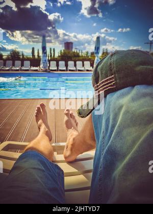 Les jambes des hommes dans un short en denim sur une chaise longue devant la piscine par temps ensoleillé. Casquette élégante sur le genou Banque D'Images