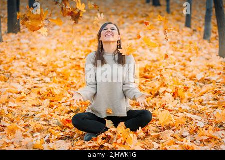 Une jeune femme souriante assise sur les feuilles s'amuser dans le parc d'automne jette les feuilles mortes Banque D'Images