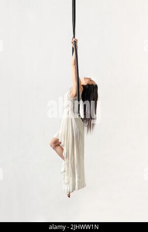 Vue latérale de la brunette pieds nus dans une longue robe suspendue sur des silks aériens tout en faisant de la gymnastique sur fond blanc Banque D'Images