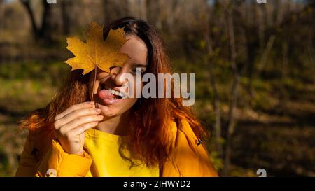 Femme caucasienne aux cheveux rouges tenant une feuille d'érable tombée.Marche d'automne. Banque D'Images