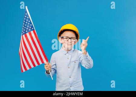 Joyeux petit garçon caucasien dans un casque de sécurité jaune tenant et agitant le drapeau américain avec le visage souriant. Futur ouvrier de la construction Banque D'Images