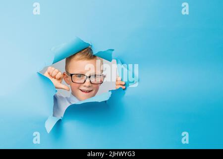 Joyeux garçon d'affaires mignon dans les lunettes de vue est s'amuser sur le mur bleu de fond, grimpe à travers un trou dans le papier. Des émotions lumineuses et drôles du garçon Banque D'Images