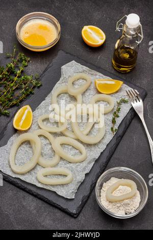 Calmar coupé dans les anneaux sur le panneau de pierre. Fourchette et huile dans la bouteille. Farine et œuf cru battu dans des bols. Pose à plat. Arrière-plan noir Banque D'Images