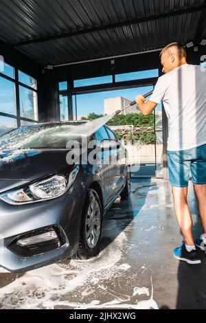 Lavage de voiture en libre-service. Un homme lave la mousse de sa voiture avec de l'eau d'un tuyau sous pression à un lavage de voiture en libre-service. Banque D'Images